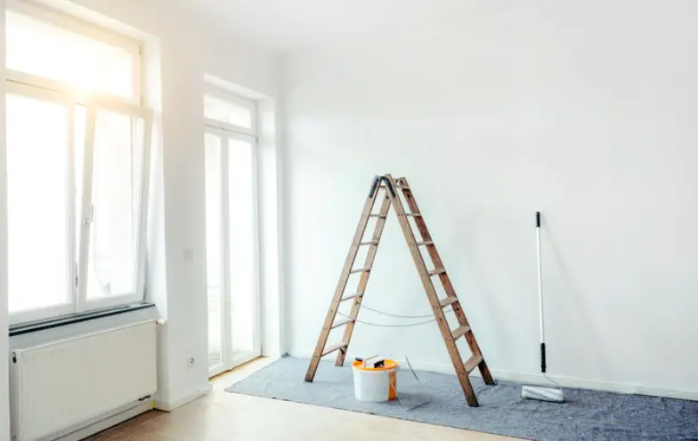 A wooden ladder stands in a bright, empty room with paint supplies on the floor.